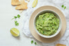 Guacamole in einer weißen Schüssel mit Untersetzer, umrundet von Limetten und Tortilla-Chips.